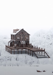 A wooden house stands on stilts surrounded by a snowy landscape, with snow-covered trees in the background. The architecture is rustic, with metal roofing and a sloped walkway leading to the entrance.