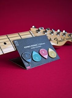 A close-up view of a guitar headstock with tuning pegs visible on the right. In front of the guitar, a display card holds three guitar picks in black, blue, pink, and yellow, labeled 'ROMBO MOSAIC | 0.65 mm MIXED COLOURS'. The background is a solid deep red.