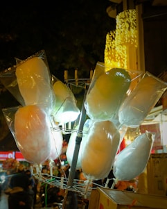A display of several bags of cotton candy illuminated by soft lights, hanging on a metal rack. The background features blurred images of a busy street or market scene with warm-toned festive lights creating a cozy ambiance.