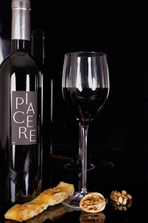 A wine bottle with a modern label is positioned next to a tall glass filled with red wine. Delicate reflections appear on the glass surface. On the table, there are a few walnut halves and a slice of bread, adding a rustic touch to the composition.