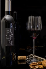 A bottle of wine labeled 'PIACERE' stands next to a filled wine glass. The setting includes wine corks, a corkscrew, and scattered walnuts on a reflective surface.