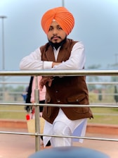 A person wearing a bright orange turban and a brown waistcoat over a white shirt is leaning against a metal railing. The background features an outdoor setting with a blurred landscape.