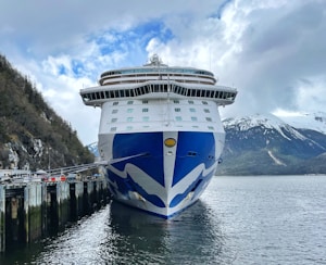 A large cruise ship is docked at a pier with a scenic backdrop of snow-capped mountains and a cloudy sky. The ship features a prominent blue and white design on its hull. The surroundings include a mix of water, greenery on the hills, and the pier structure.