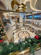 A luxurious, multi-story interior with festive holiday decorations, including a large Christmas tree, ornate railings, and opulent lighting fixtures. The floors are polished with an intricate design, and there are seating areas with people scattered around.