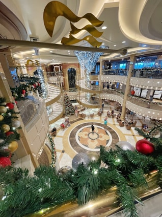 A luxurious, multi-story interior with festive holiday decorations, including a large Christmas tree, ornate railings, and opulent lighting fixtures. The floors are polished with an intricate design, and there are seating areas with people scattered around.