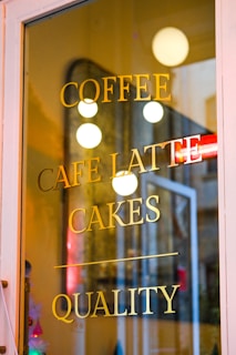 A glass door with the words 'COFFEE', 'CAFE LATTE', 'CAKES', and 'QUALITY' printed in gold lettering. The reflection of round lights and an open door can be seen on the glass surface.