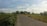 A rural road stretches through a landscape with green fields on either side, lined with trees and bushes. Telephone poles and power lines run parallel to the road, leading toward the distant horizon. The sky is mostly clear with some scattered clouds.