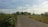 A rural road stretches through a landscape with green fields on either side, lined with trees and bushes. Telephone poles and power lines run parallel to the road, leading toward the distant horizon. The sky is mostly clear with some scattered clouds.