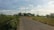 A rural road stretches through a landscape with green fields on either side, lined with trees and bushes. Telephone poles and power lines run parallel to the road, leading toward the distant horizon. The sky is mostly clear with some scattered clouds.
