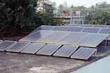 A collection of solar panels is installed on the flat rooftop of a building. Surrounding the roof, there are trees and additional buildings visible in the background. Clothes hang drying on a line, adding a touch of daily life to the scene.