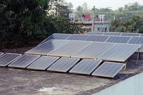 A collection of solar panels is installed on the flat rooftop of a building. Surrounding the roof, there are trees and additional buildings visible in the background. Clothes hang drying on a line, adding a touch of daily life to the scene.