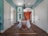 A hallway with teal walls and white doors, leading to a room with an orange accent wall. A wooden chair sits at a table covered in a blue and white patterned tablecloth over a decorative rug. A black chandelier hangs from the ceiling, and greenery is visible outside the tall windows.