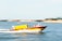 A fast-moving yellow and red emergency boat with 'Ambulanza' written on the side cuts through the water. Two people are visible on board, with blurred water and background indicating motion.