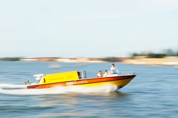 A fast-moving yellow and red emergency boat with 'Ambulanza' written on the side cuts through the water. Two people are visible on board, with blurred water and background indicating motion.