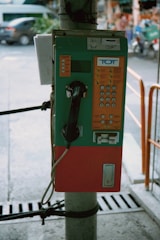 A vintage public payphone is mounted on a pole, featuring a green and orange casing. The keypad and coin slots are visible, and the phone hangs on the left side. The surrounding environment suggests an outdoor urban setting with a blurred street and vehicles in the background.