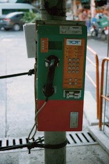 A vintage public payphone is mounted on a pole, featuring a green and orange casing. The keypad and coin slots are visible, and the phone hangs on the left side. The surrounding environment suggests an outdoor urban setting with a blurred street and vehicles in the background.