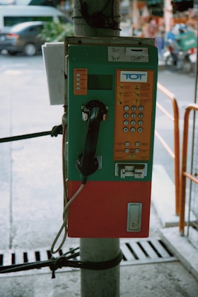 A vintage public payphone is mounted on a pole, featuring a green and orange casing. The keypad and coin slots are visible, and the phone hangs on the left side. The surrounding environment suggests an outdoor urban setting with a blurred street and vehicles in the background.