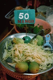 A metal bowl contains shredded mango and whole mangoes. A blue sign with '50 MANGO' is placed in the bowl. Nearby are leafy greens in a clay pot and a blue vegetable peeler.