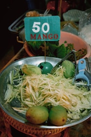A metal bowl contains shredded mango and whole mangoes. A blue sign with '50 MANGO' is placed in the bowl. Nearby are leafy greens in a clay pot and a blue vegetable peeler.
