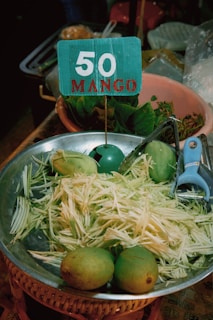 A metal bowl contains shredded mango and whole mangoes. A blue sign with '50 MANGO' is placed in the bowl. Nearby are leafy greens in a clay pot and a blue vegetable peeler.