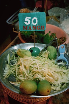 A metal bowl contains shredded mango and whole mangoes. A blue sign with '50 MANGO' is placed in the bowl. Nearby are leafy greens in a clay pot and a blue vegetable peeler.