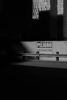 A corrugated metal barrier with a warning sign in Spanish, reading 'PELIGRO EXCAVACIÓN,' is partially illuminated by light. Large, shadowed buildings with a grid pattern of windows loom in the background, creating a dramatic play of light and shadow.