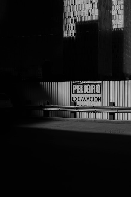 A corrugated metal barrier with a warning sign in Spanish, reading 'PELIGRO EXCAVACIÓN,' is partially illuminated by light. Large, shadowed buildings with a grid pattern of windows loom in the background, creating a dramatic play of light and shadow.