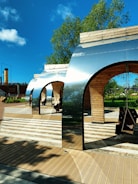 Several modern, metallic and wooden arch structures with swings are situated in an outdoor area under a clear blue sky. The ground features a patterned wooden and tile surface, and there are people sitting on the swings. Green trees and shrubs are visible in the background, contributing to a serene and pleasant environment.