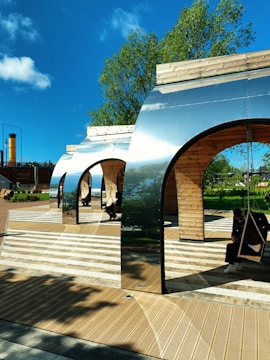 Several modern, metallic and wooden arch structures with swings are situated in an outdoor area under a clear blue sky. The ground features a patterned wooden and tile surface, and there are people sitting on the swings. Green trees and shrubs are visible in the background, contributing to a serene and pleasant environment.