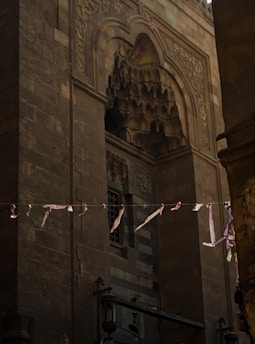 An ornate stone facade features intricate architectural details and carvings, with an arched design at the top. Faded pink ribbons are strung across, adding a touch of contrast to the muted color palette of the stone. The facade is part of a larger structure with elements of historical and cultural significance.