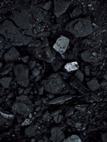 A close-up view of a surface covered in dark, rough asphalt with scattered pieces of white rubble and debris. The texture is uneven and consists of various-sized stones and broken concrete fragments.