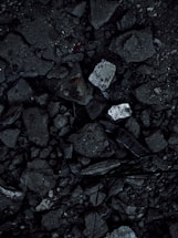 A close-up view of a surface covered in dark, rough asphalt with scattered pieces of white rubble and debris. The texture is uneven and consists of various-sized stones and broken concrete fragments.