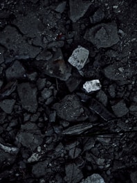 A close-up view of a surface covered in dark, rough asphalt with scattered pieces of white rubble and debris. The texture is uneven and consists of various-sized stones and broken concrete fragments.