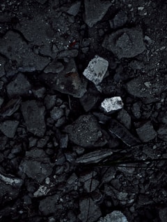 A close-up view of a surface covered in dark, rough asphalt with scattered pieces of white rubble and debris. The texture is uneven and consists of various-sized stones and broken concrete fragments.