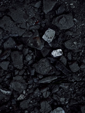A close-up view of a surface covered in dark, rough asphalt with scattered pieces of white rubble and debris. The texture is uneven and consists of various-sized stones and broken concrete fragments.