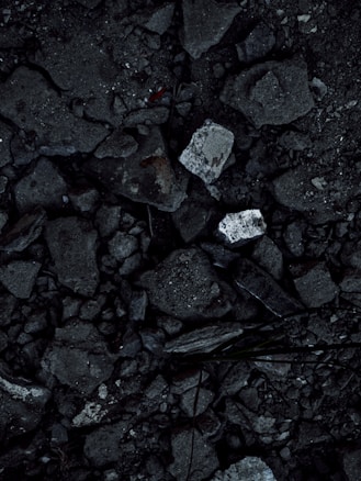 A close-up view of a surface covered in dark, rough asphalt with scattered pieces of white rubble and debris. The texture is uneven and consists of various-sized stones and broken concrete fragments.