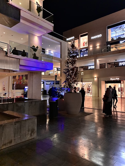 An outdoor shopping area at night, featuring multiple illuminated retail store fronts with the name 'Mango' visible. There is a decorative tree with lights and large planters with greenery. People can be seen walking and standing around the area, and the ground appears wet, perhaps from recent rain. The architecture includes multiple levels with glass railings and a modern design.
