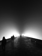 A foggy scene with several people walking on a stone pathway illuminated by bright lights. The mist creates a mysterious and surreal atmosphere, with silhouettes of pedestrians gradually fading into the light.