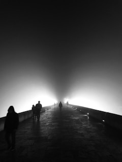 A foggy scene with several people walking on a stone pathway illuminated by bright lights. The mist creates a mysterious and surreal atmosphere, with silhouettes of pedestrians gradually fading into the light.