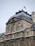 Ornate architectural details on a historic building featuring sculptures and intricate carvings. The structure is topped with a dark, steep roof, and a French flag flutters at the peak. The facade showcases symmetrical windows and detailed stonework.