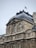 Ornate architectural details on a historic building featuring sculptures and intricate carvings. The structure is topped with a dark, steep roof, and a French flag flutters at the peak. The facade showcases symmetrical windows and detailed stonework.