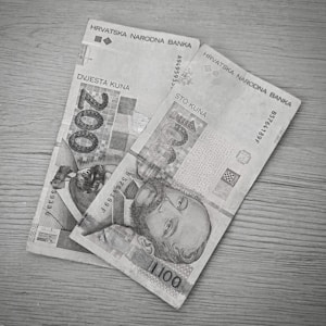Two paper banknotes, a 200 kuna bill and a 100 kuna bill, are placed on a wooden surface. The bills are from the Croatian National Bank and feature portraits of historical figures along with text and intricate designs.