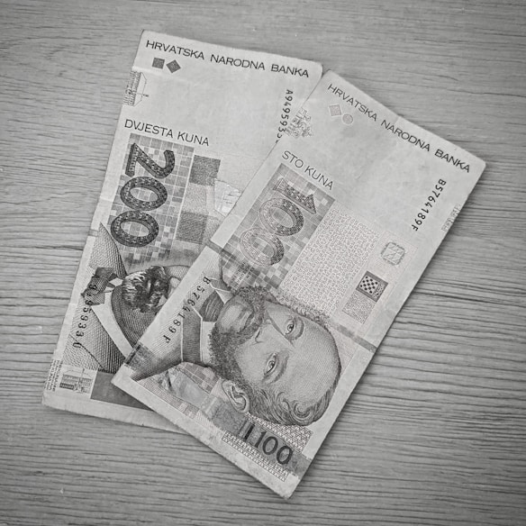 Two paper banknotes, a 200 kuna bill and a 100 kuna bill, are placed on a wooden surface. The bills are from the Croatian National Bank and feature portraits of historical figures along with text and intricate designs.