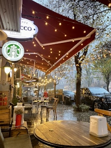 A cozy outdoor caf&eacute; setting with string lights hanging under a red canopy. Wooden tables and metal chairs are arranged along a wet pavement, suggesting recent rain. Trees line the sidewalk, adding a natural element to the urban environment. A person dressed in red and holding an umbrella walks in the background, contributing to the busy, yet intimate atmosphere.