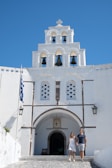 A couple is walking hand in hand in front of a white-washed building with a bell tower featuring several arches and bells. The sky is clear with a vibrant blue color, and a Greek flag is visible on the left side. The architecture has traditional Greek features.