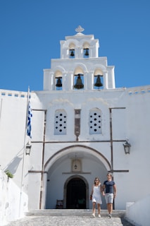 A couple is walking hand in hand in front of a white-washed building with a bell tower featuring several arches and bells. The sky is clear with a vibrant blue color, and a Greek flag is visible on the left side. The architecture has traditional Greek features.