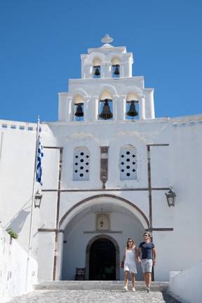 A couple is walking hand in hand in front of a white-washed building with a bell tower featuring several arches and bells. The sky is clear with a vibrant blue color, and a Greek flag is visible on the left side. The architecture has traditional Greek features.