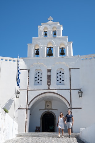 A couple is walking hand in hand in front of a white-washed building with a bell tower featuring several arches and bells. The sky is clear with a vibrant blue color, and a Greek flag is visible on the left side. The architecture has traditional Greek features.