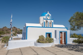 A white building with blue accents and a domed roof stands prominently under a clear blue sky. The structure features three bells in a small tower and is surrounded by greenery. A Greek flag is flying on a pole nearby. The setting is peaceful and sunny, with distant hills visible in the background.