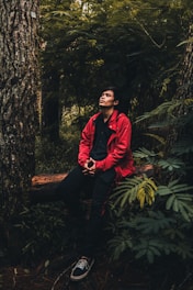 A person in a red jacket sits contemplatively in a lush forest setting, surrounded by tall trees and abundant greenery. The person's gaze is directed upwards, adding to the introspective atmosphere.